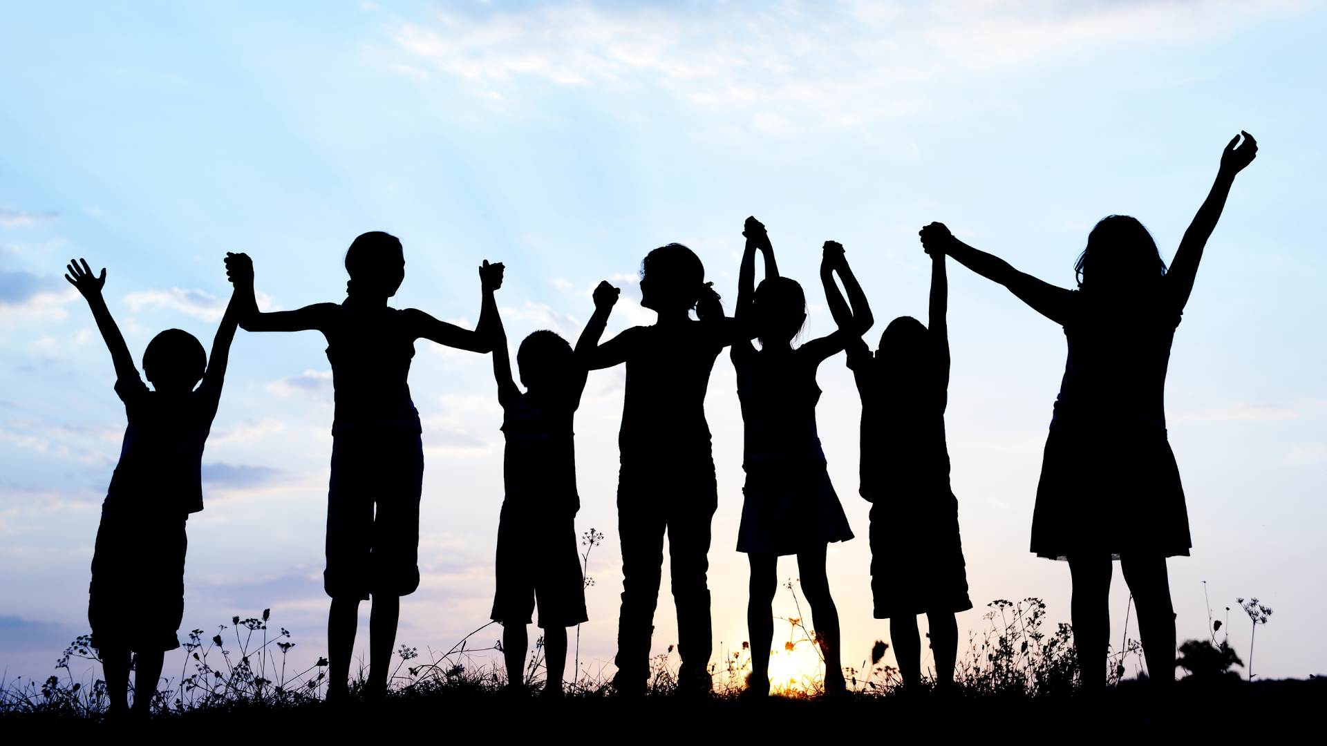 Silhouetten von Kindern, die Hände fassend in einer Reihe stehen und ihre Hände in die Luft jubelnd werfen vor einem Sonnenuntergang
